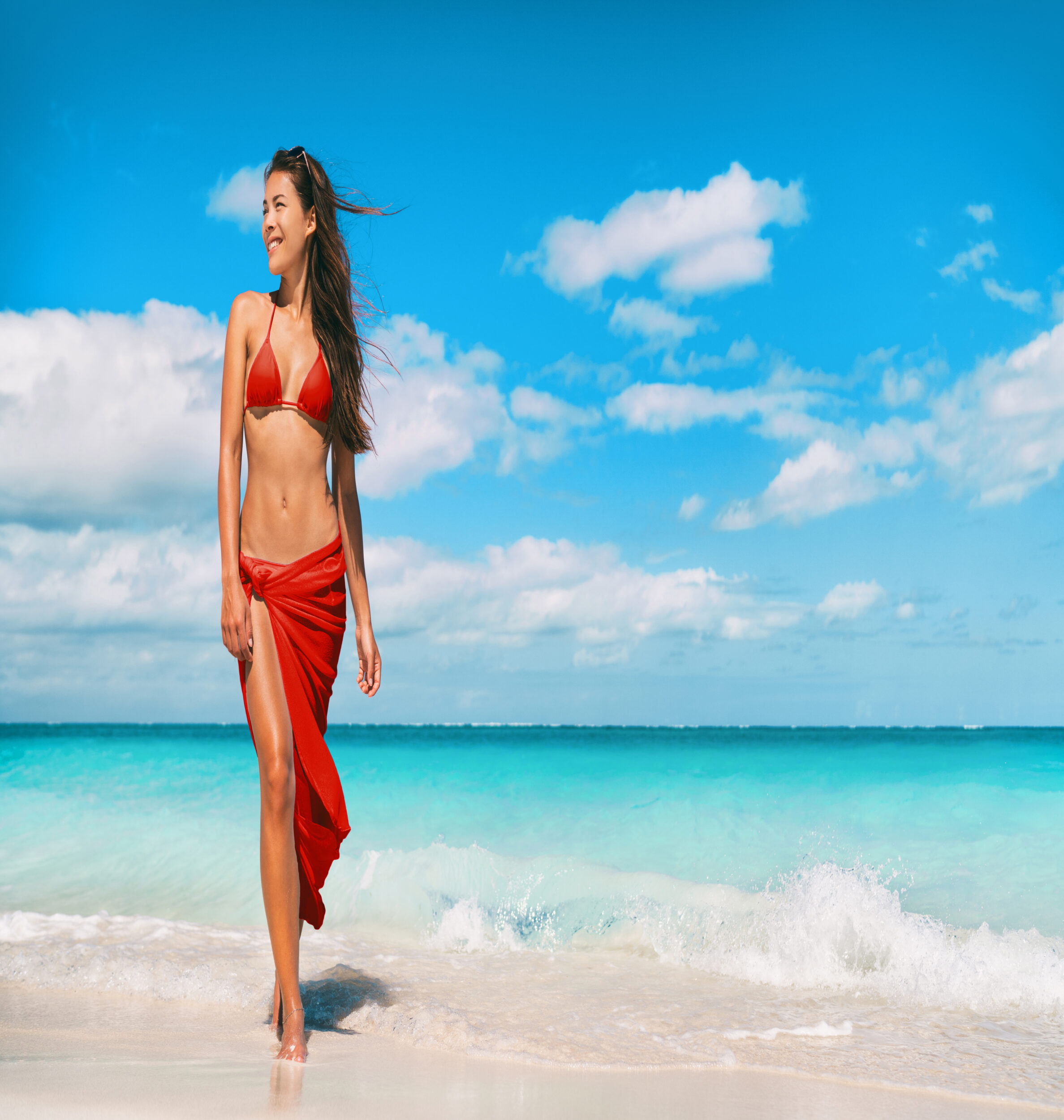 Woman, wearing a red bikini top and stylish sarong, looks into the distance smiling, feeling happy with her toned tummy after surgery